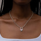 A close-up of a model wearing a silver chain necklace with a small pendant featuring a pink heart and a tiny red gemstone, paired with a white ribbed tank top.