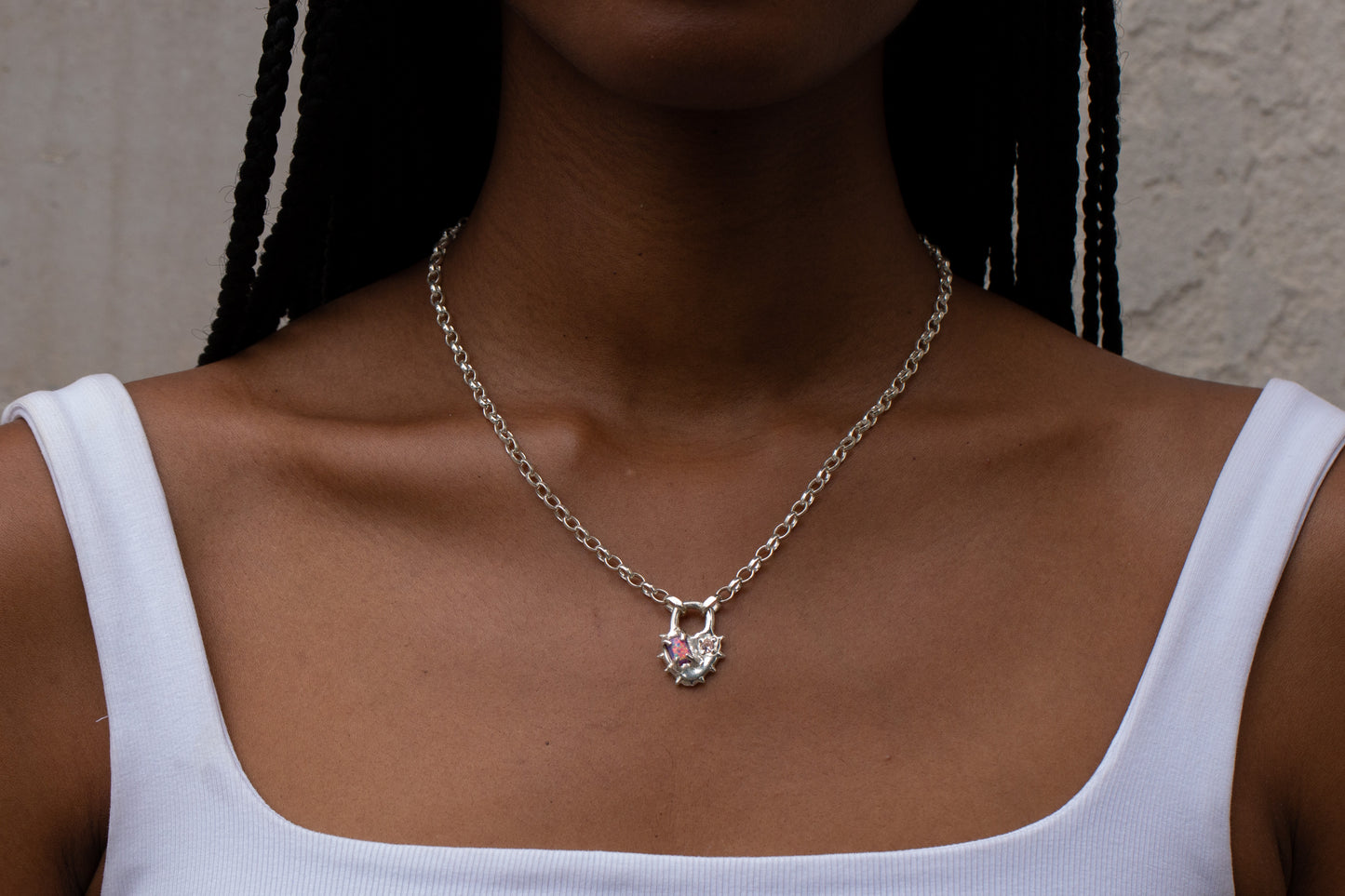 A close-up of a model wearing a silver chain necklace with a small pendant featuring a pink heart and a tiny red gemstone, paired with a white ribbed tank top.